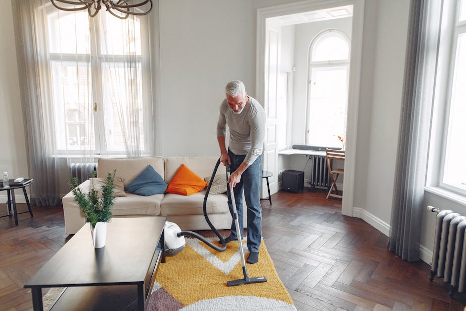 A person wearing casual clothing and beige shoes is using a vacuum cleaner with a flexible hose attachment to clean a traditional patterned rug with floral motifs in shades of beige, green, and orange